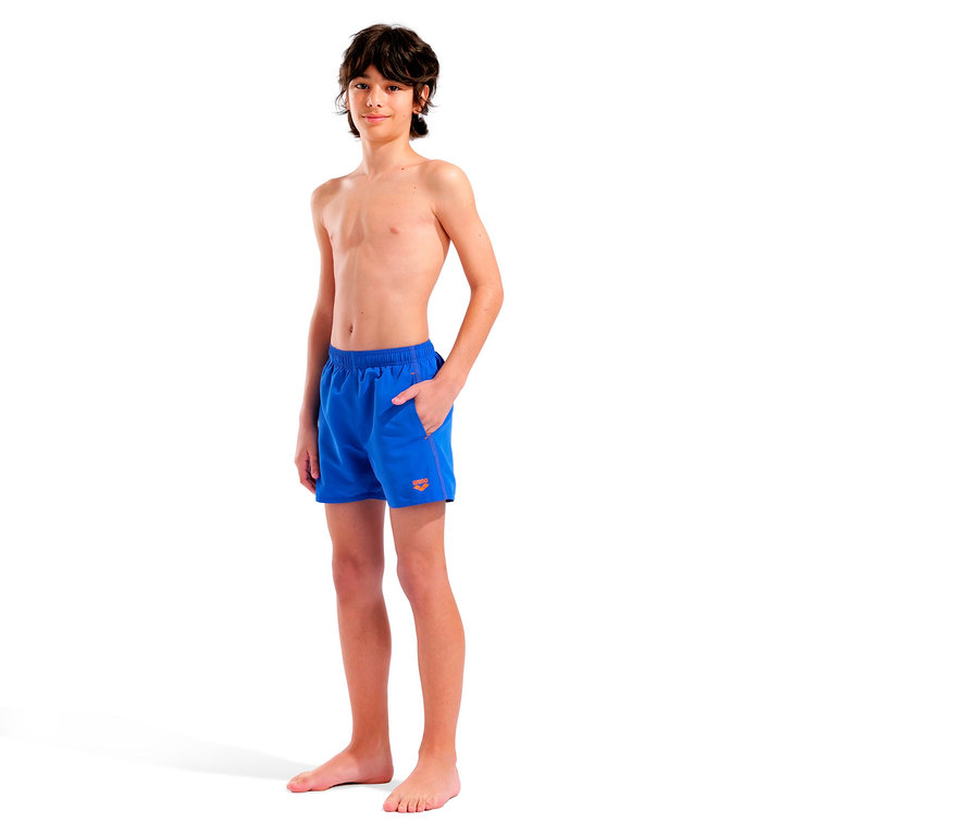 Młody, bosy chłopak z brązowymi włosami stoi na białym tle w niebieskich szortach kąpielowych z pomarańczowym logo.