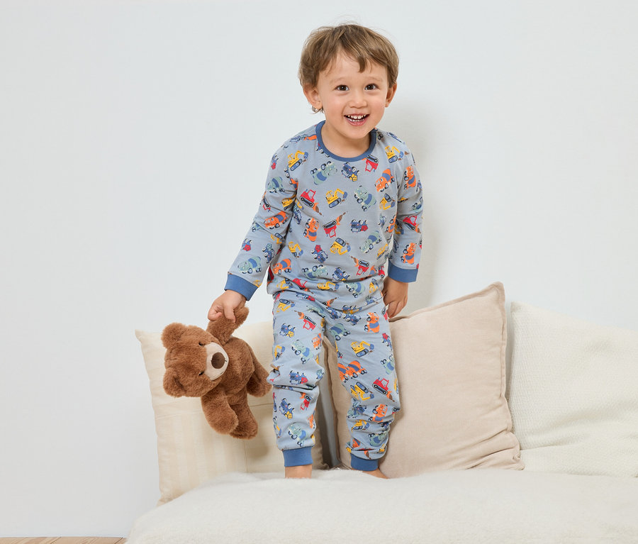 Uśmiechnięty chłopiec w niebieskiej piżamie z wzorem pojazdów budowlanych stoi z pluszowym misiem na sofie.