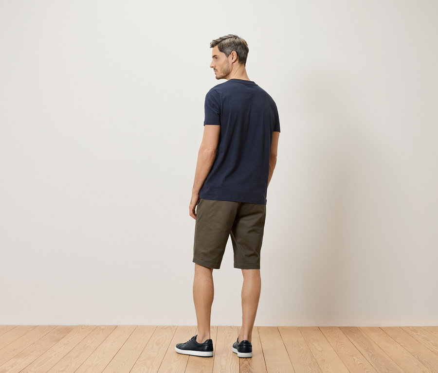 Mężczyzna stoi tyłem do kamery, ubrany w granatowy T-shirt, khaki szorty i granatowe buty.