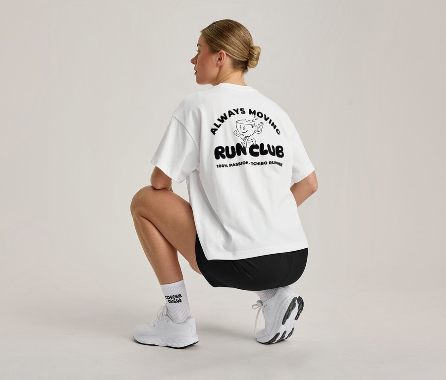 Kobieta z blond kokiem kuca, widziana od tyłu. Ma na sobie białą, oversize'ową koszulkę sportową z czarną grafiką 'ALWAYS MOVING RUN CLUB' na plecach, czarne szorty, białe skarpetki i białe buty sportowe.