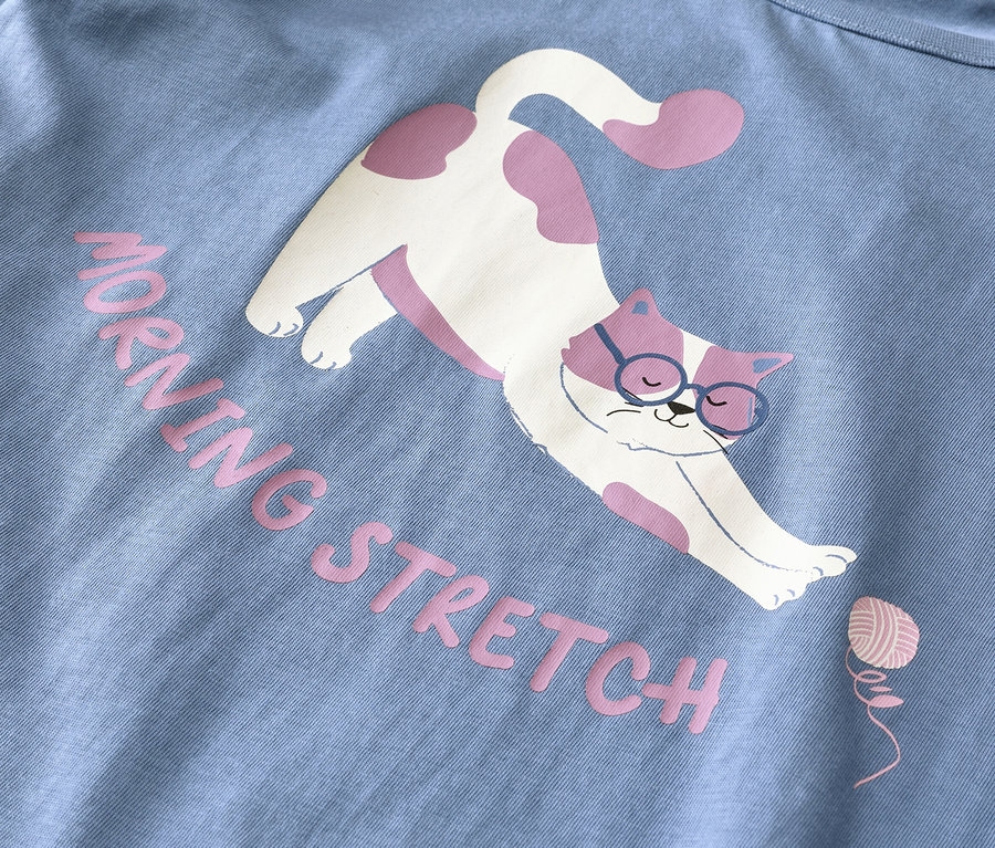 Jasnoniebieski materiał z nadrukiem białego kota w różowe cętki i niebieskich okularach, który się przeciąga. Różowy napis głosi "MORNING STRETCH" obok małego różowego kłębka włóczki.