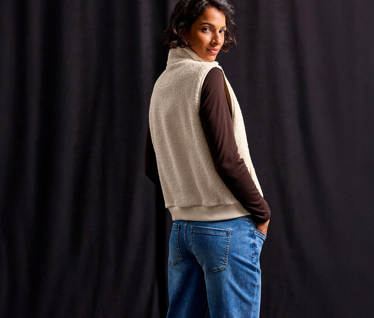Kobieta w brązowej bluzce i niebieskich dżinsach, ubrana w pluszową kamizelkę od Street One, patrzy przez ramię.