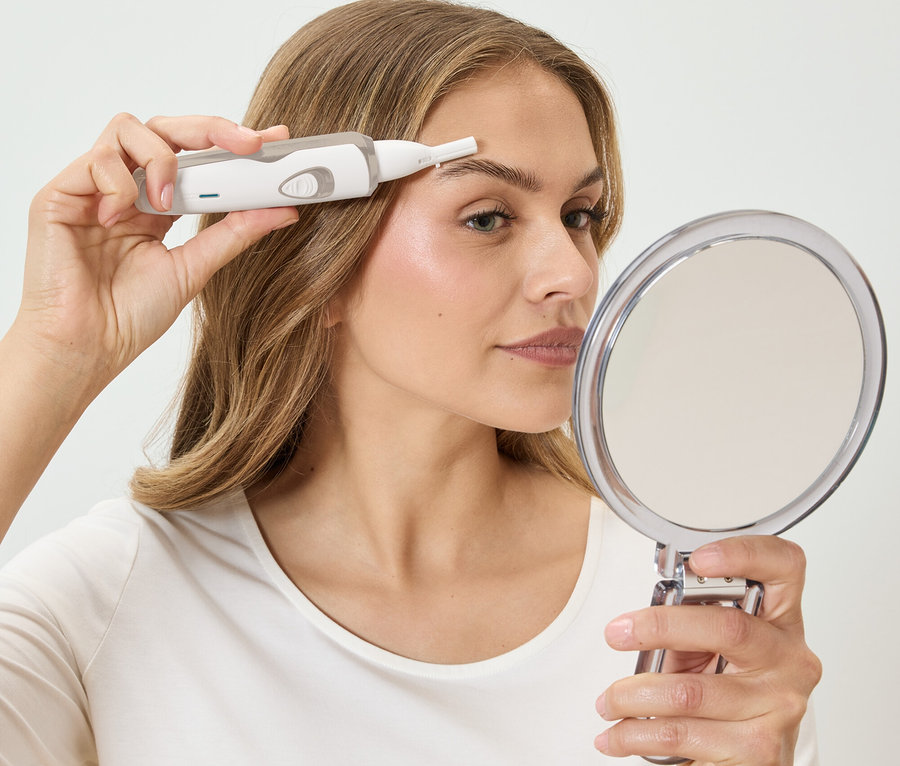 Kobieta reguluje brwi białym depilatorem, patrząc w okrągłe lustro.