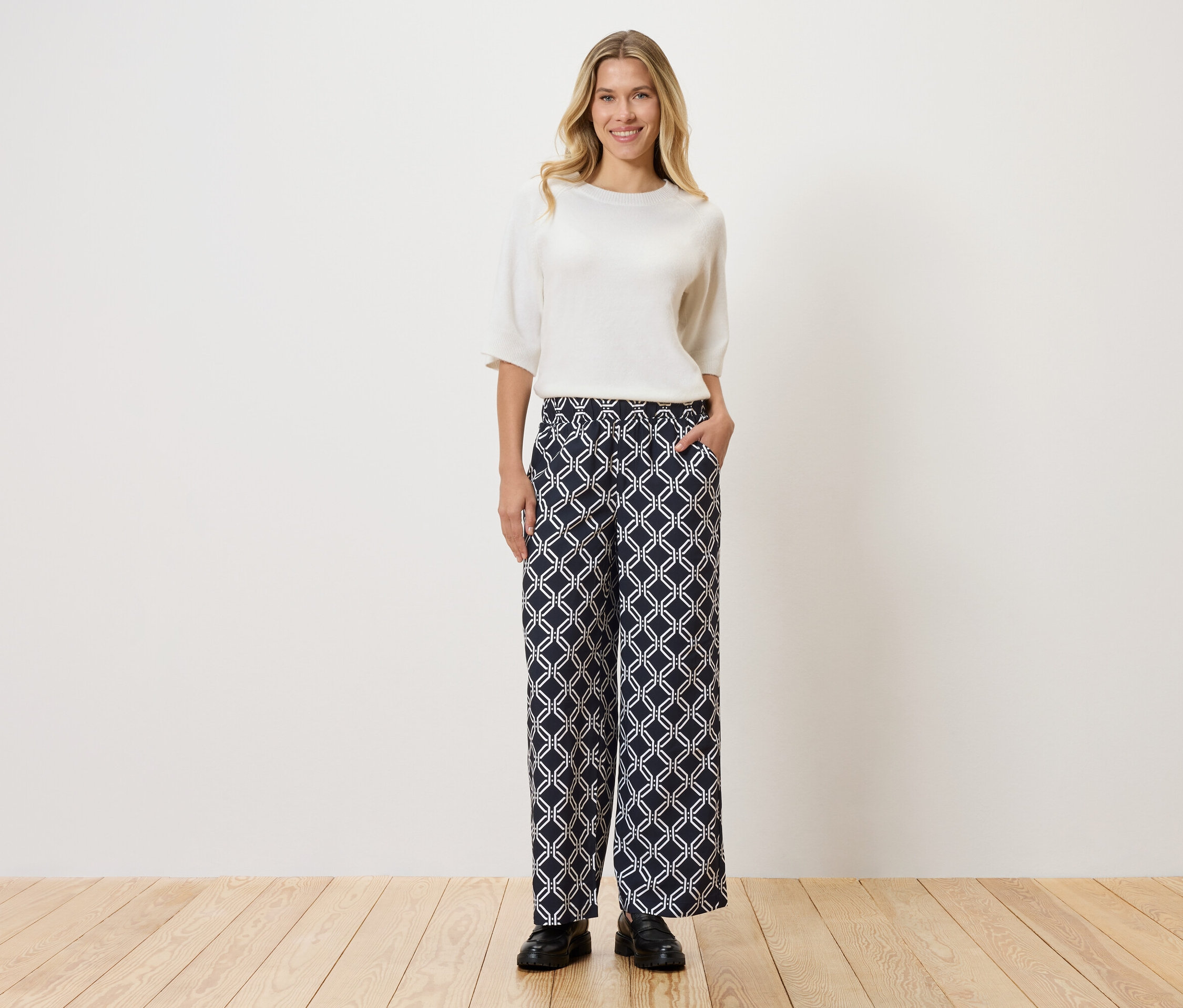 Kobieta w białym topie i wzorzystych spodniach stojąca na drewnianej podłodze przed ścianą.