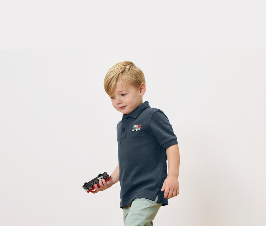 Blondwłosy chłopiec trzyma w ręku zabawkę-autko, ubrany w granatową koszulkę polo z aplikacją małego pociągu.