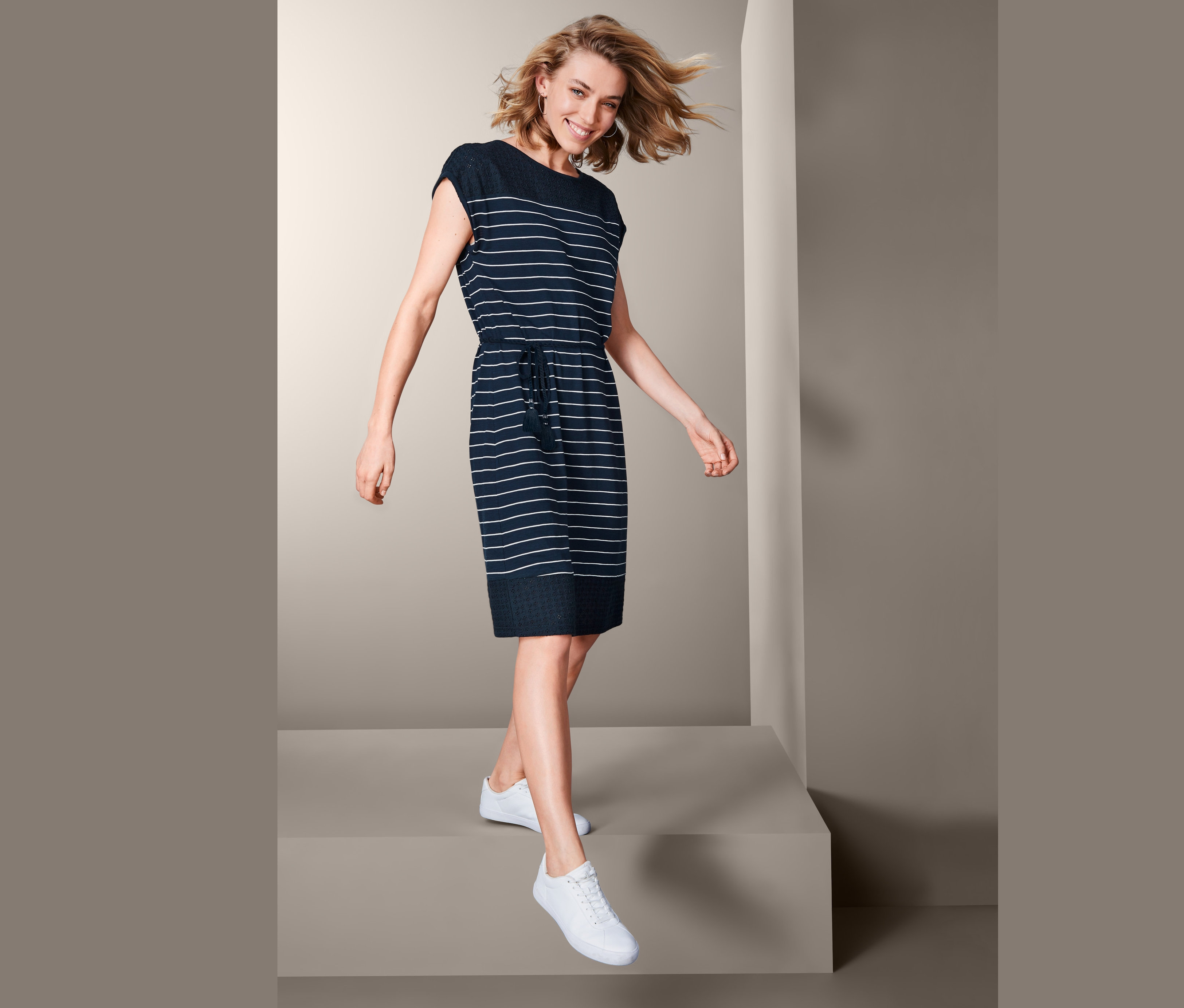 Kobieta idzie, ubrana w sukienkę z dżerseju z ażurowym haftem i białe tenisówki.