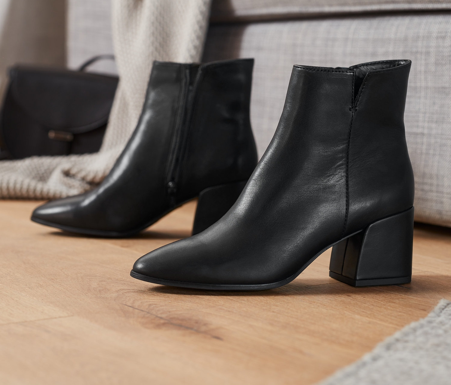 Czarne skórzane botki na drewnianej podłodze obok szarej kanapy.