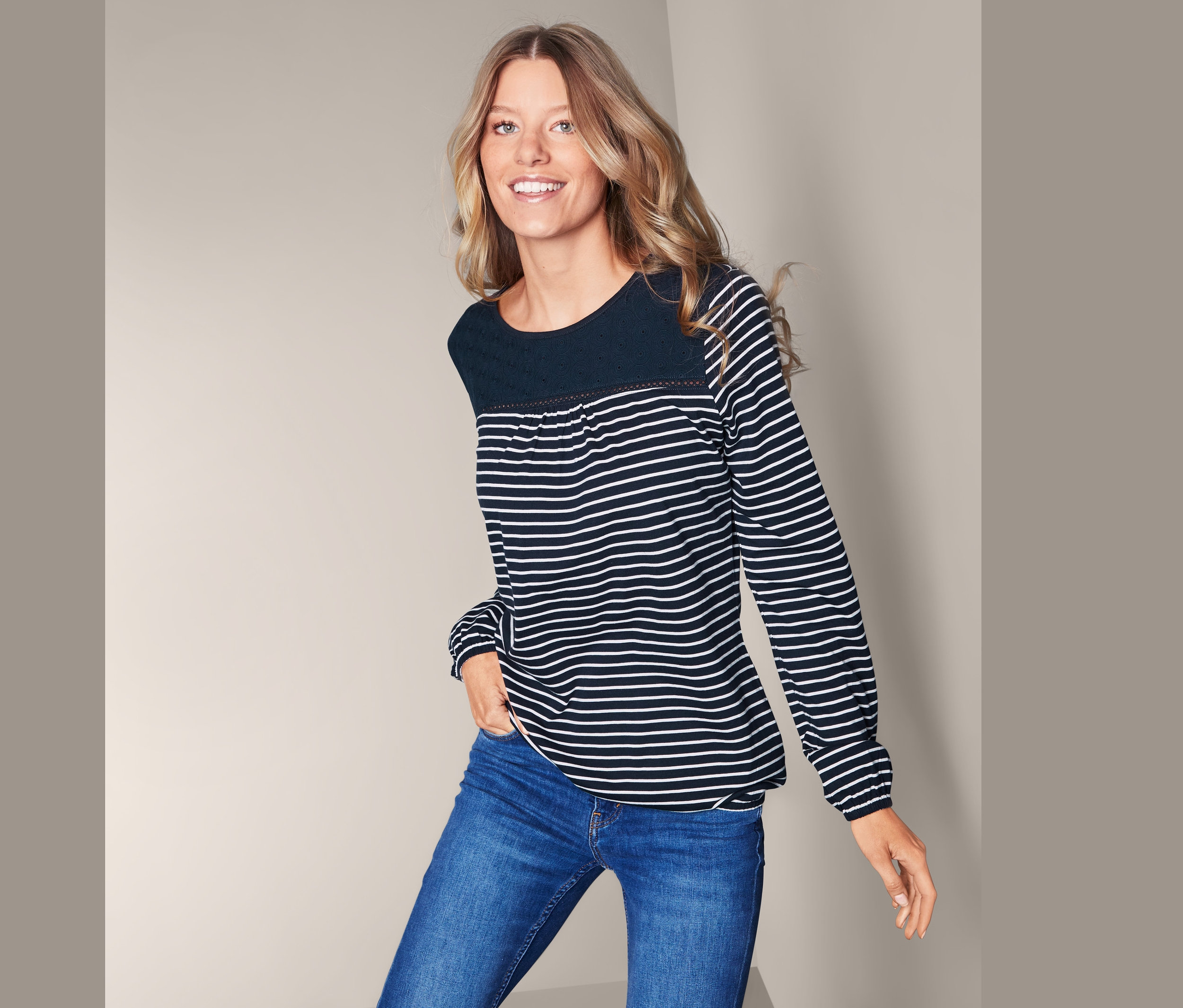 Kobieta pozuje w bluzce damskiej z haftem w niebiesko-białe paski i niebieskich dżinsach.