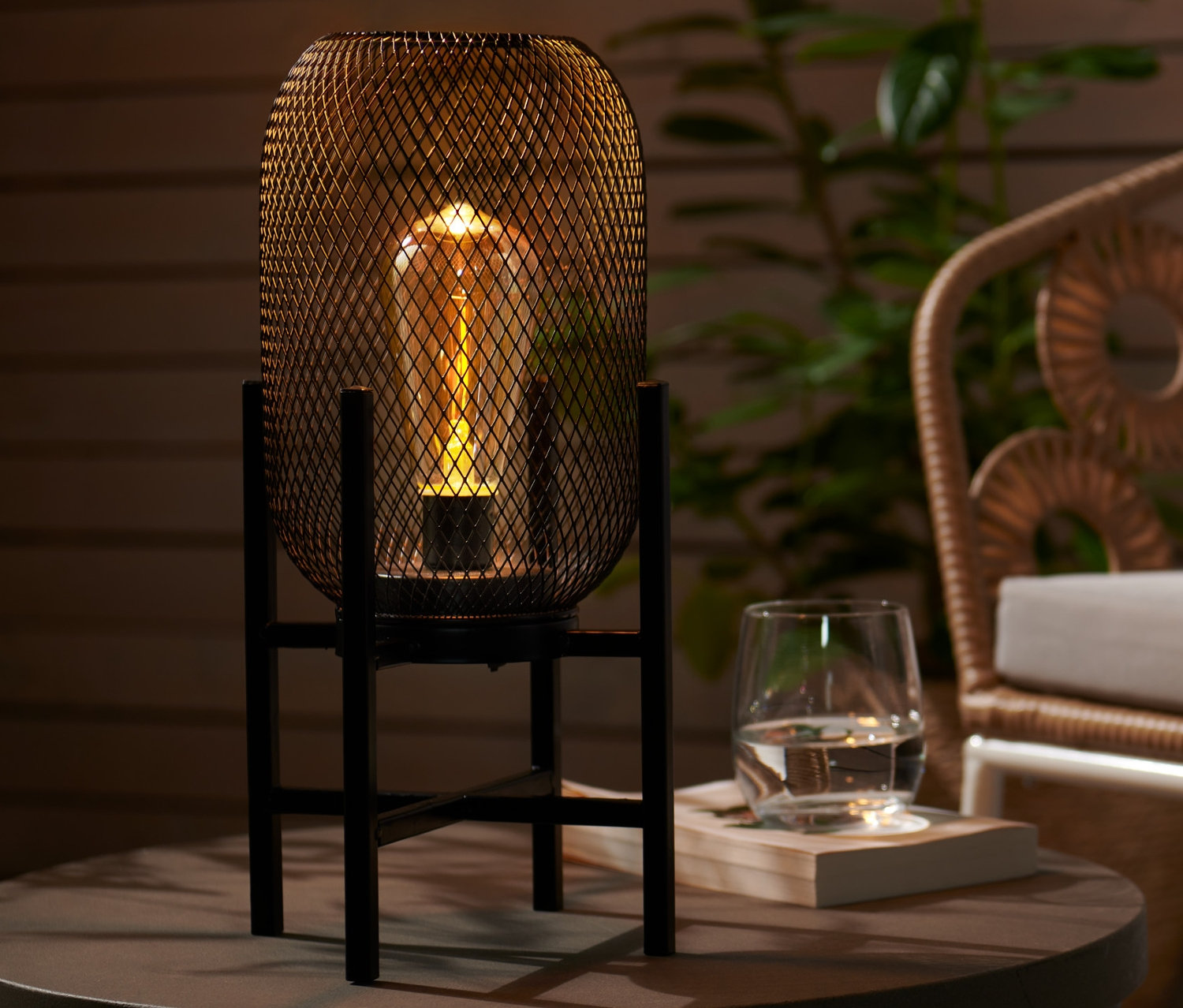 Plenerowa lampka stołowa LED stoi na okrągłym stole obok szklanki wody. W tle widać rattanowy fotel.