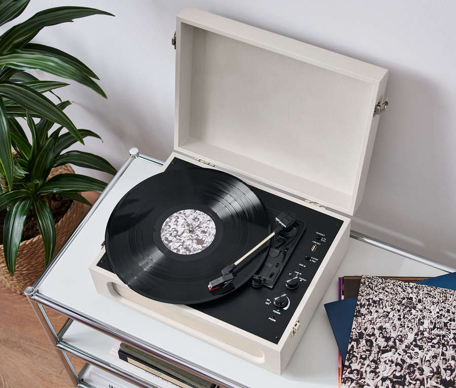 Otwarty, przenośny gramofon retro z funkcją Bluetooth® stoi na białym stole i odtwarza płytę. Obok leżą płyty winylowe, w tle roślina w koszu.