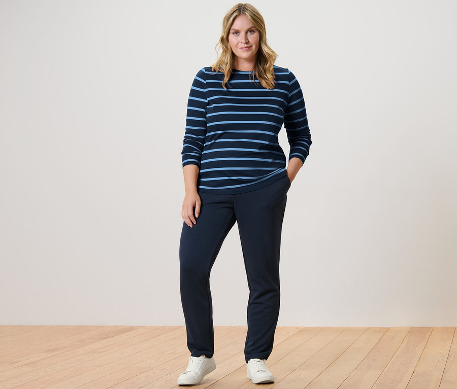 Kobieta stoi w granatowych spodniach dresowych i pasiastej koszulce z długimi rękawami.