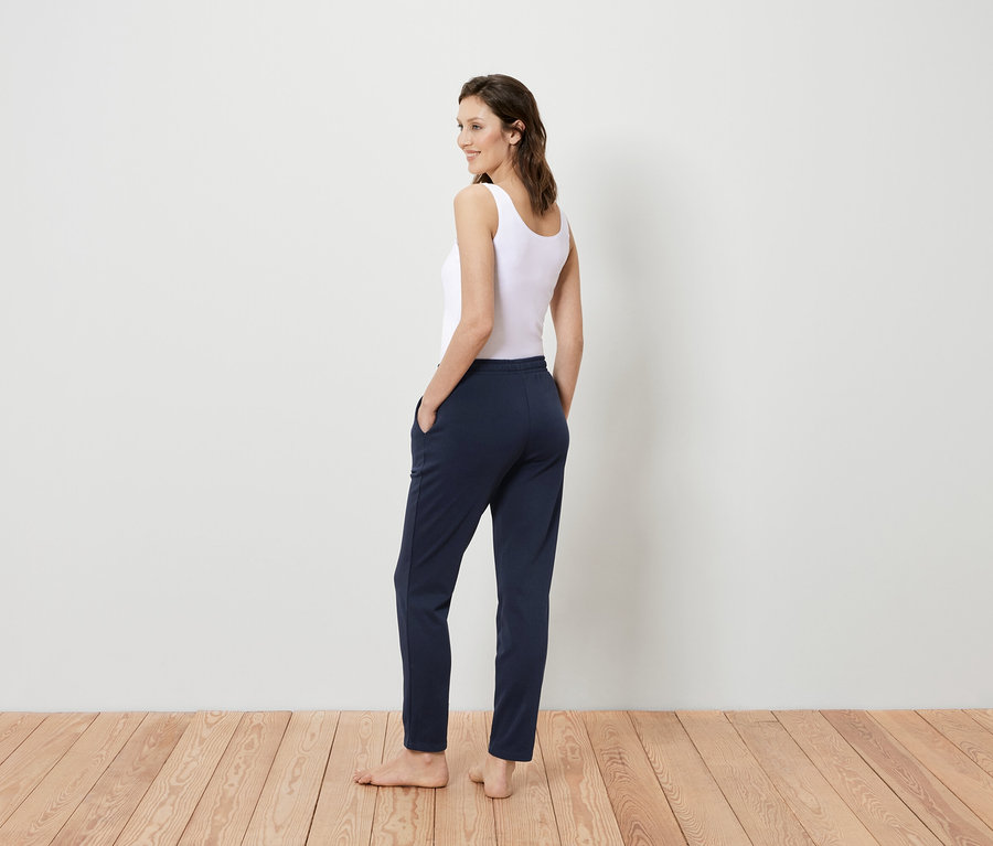 Kobieta w białym podkoszulku i granatowych spodniach stoi tyłem do kamery, spoglądając przez ramię.