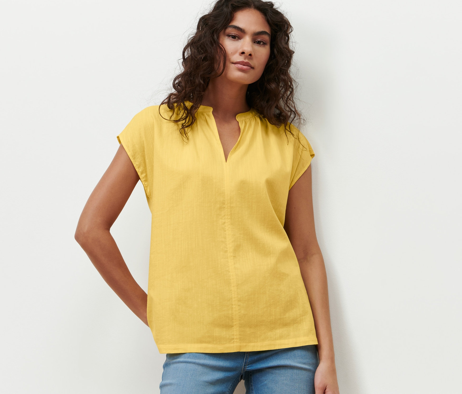 Kobieta pozuje w żółtej bluzce z krótkim rękawem i niebieskich dżinsach.