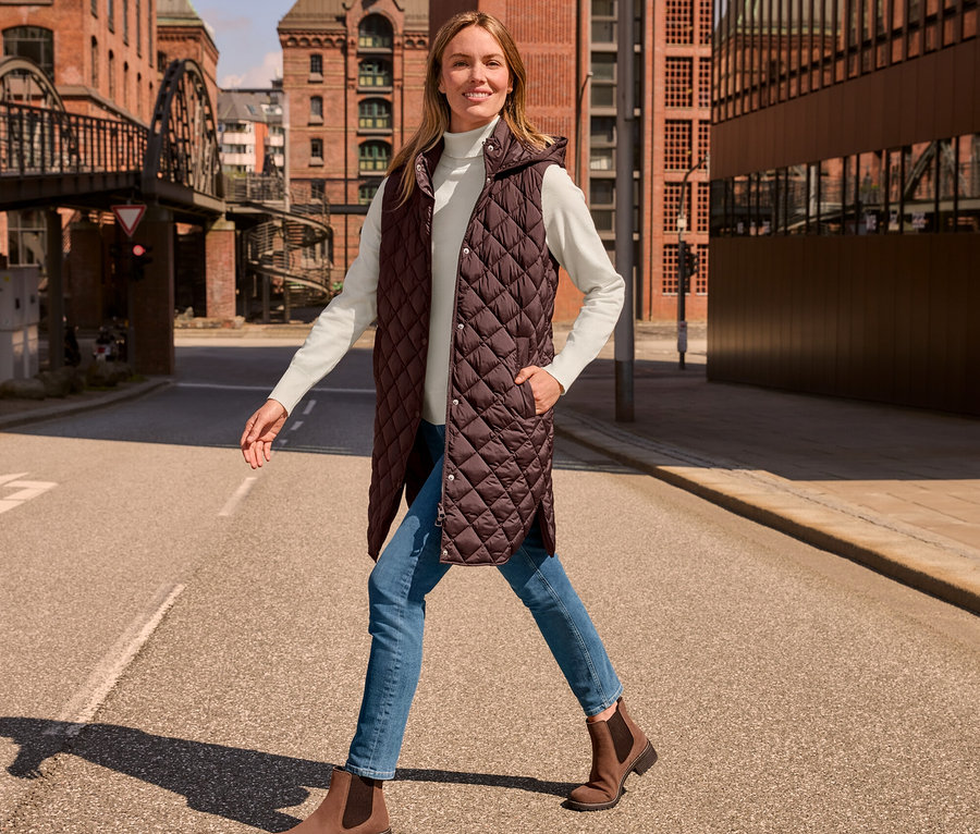 Kobieta idzie ulicą w długiej kamizelce pikowanej z kapturem, kremowobiałym golfie z cienkiej dzianiny, jeansach o wąskim kroju – fason „Emma”, niebieskich i zamszowych botkach.