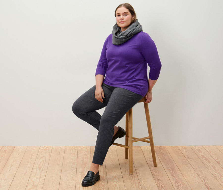 Kobieta siedzi na stołku ubrana w spodnie stretchowe i fioletową koszulkę z rękawami o długości 3/4.