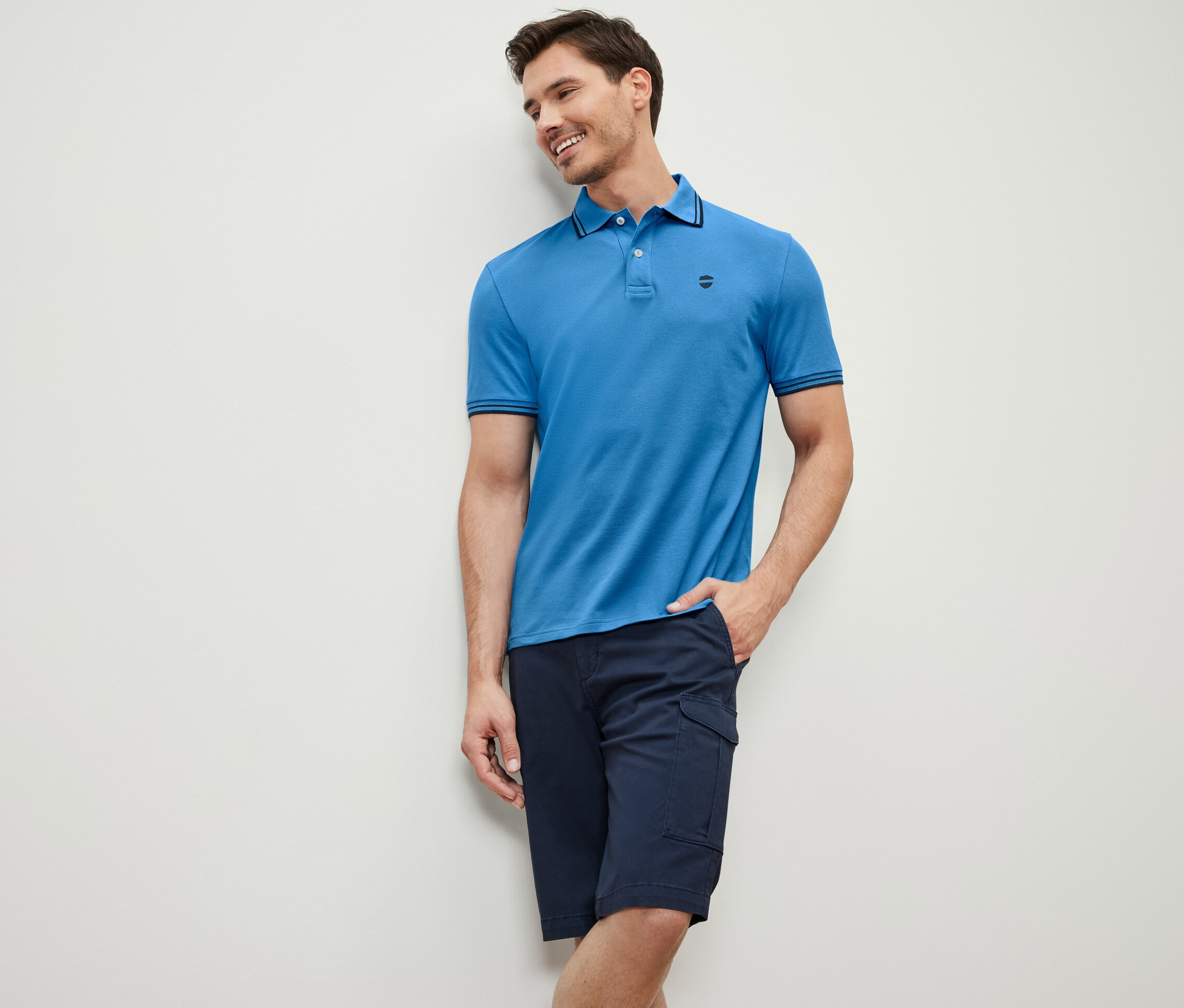 Mężczyzna oparty o ścianę ma na sobie niebieską koszulkę polo o splocie piqué i granatowe szorty.