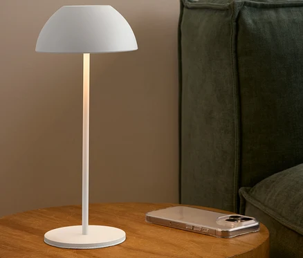Biała lampa stoi na drewnianym stoliku obok telefonu komórkowego i zielonej sofy.