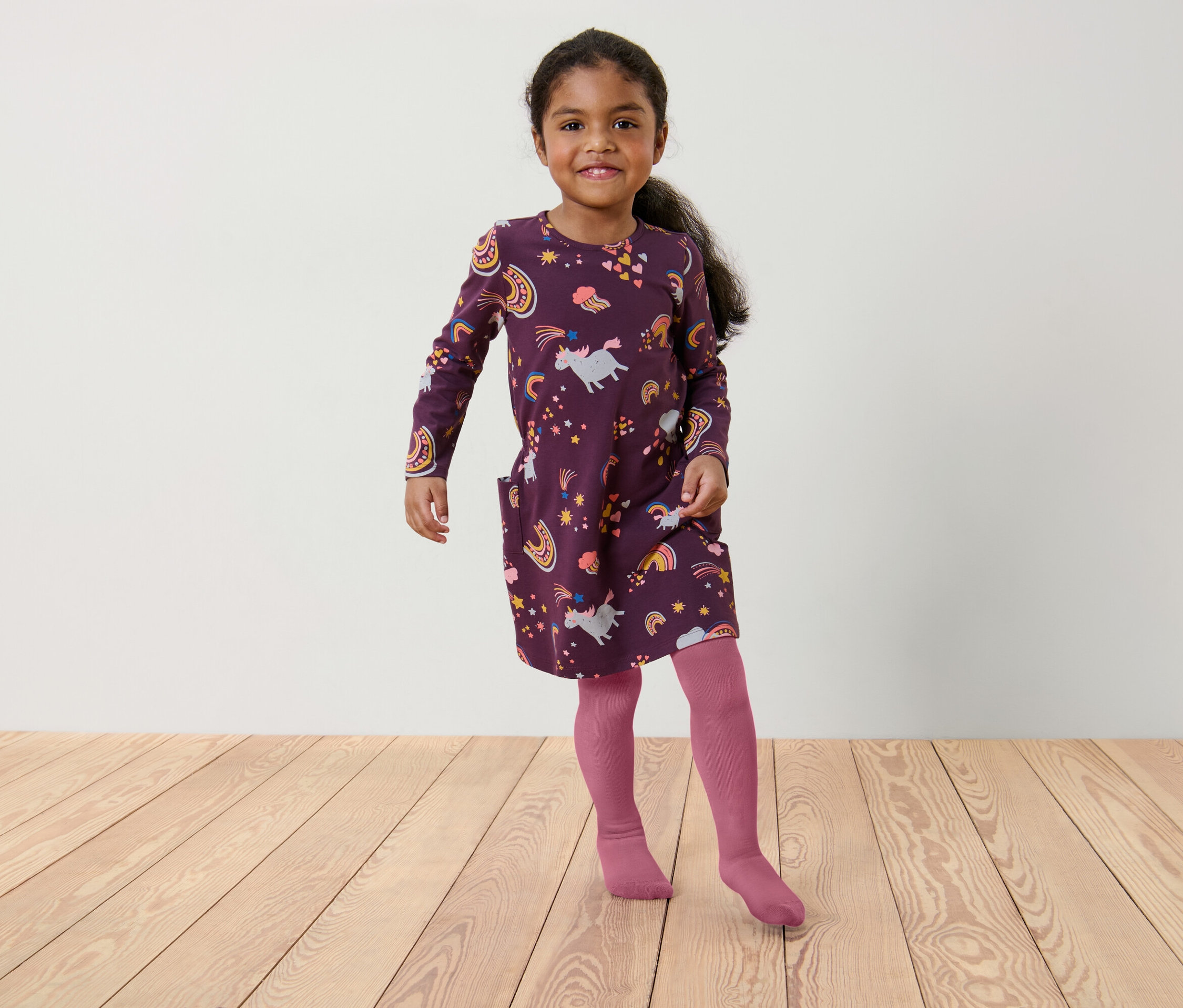 Dziewczynka w bordowej sukience z jednorożcami i tęczami oraz różowych rajstopach stoi na drewnianej podłodze.