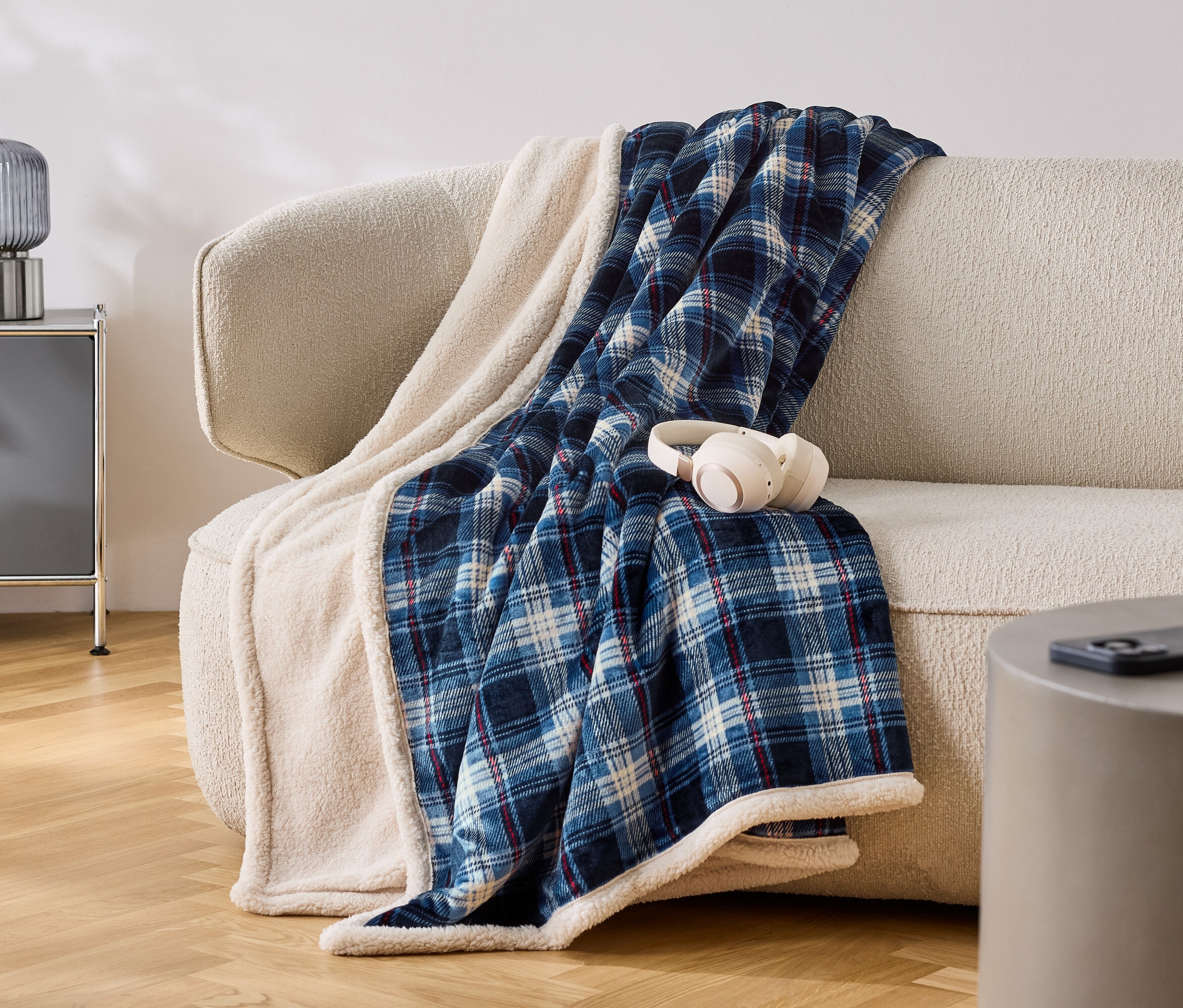 Na beżowej sofie leży niebiesko-biały koc w kratę z białym obszyciem. Na kocu leżą białe słuchawki.