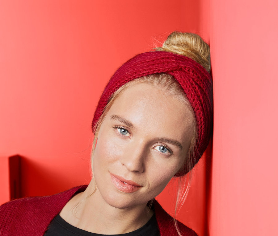 Blondynka opiera się o koralowo-czerwoną ścianę, ma na sobie opaskę na czoło z dzianiny.