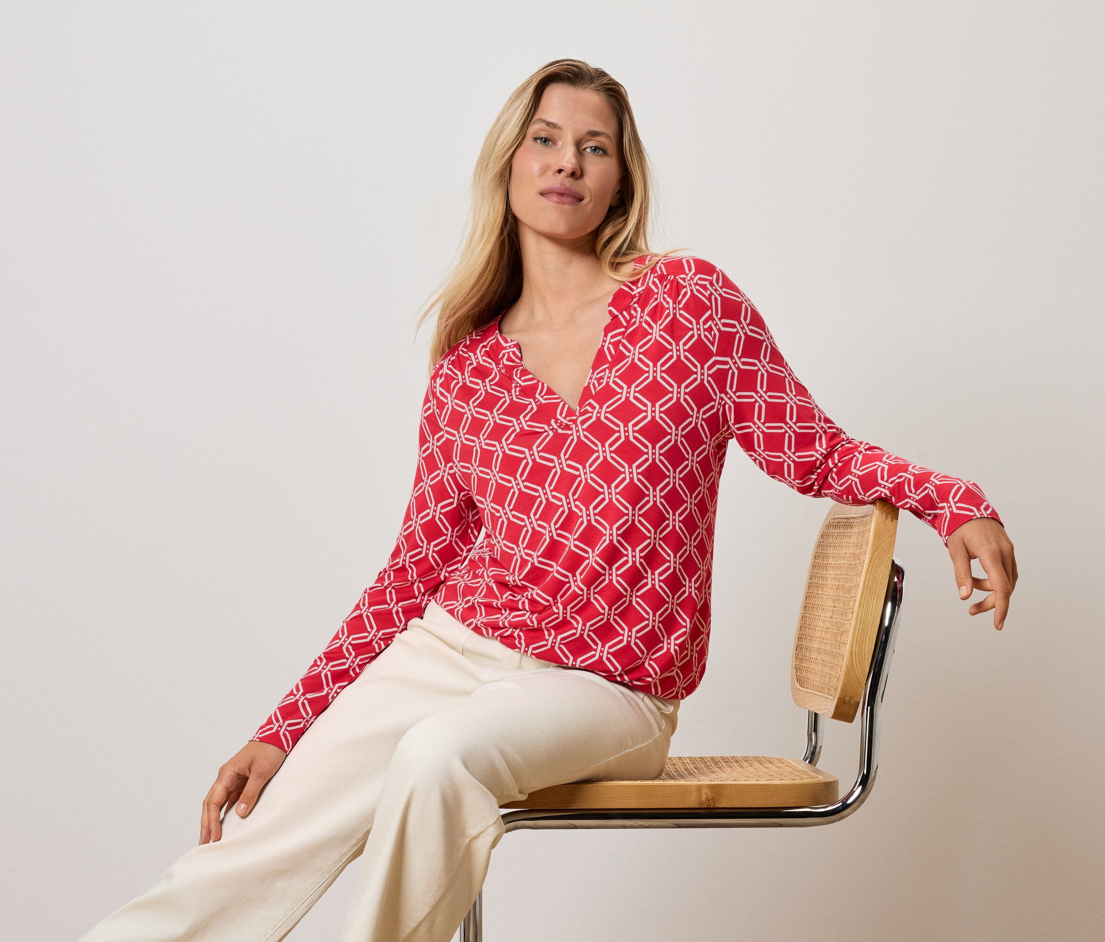 Blondynka siedzi na krześle, ubrana w czerwono-białą bluzkę i kremowe spodnie.