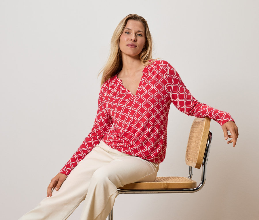 Blondynka siedzi na krześle, ubrana w czerwono-białą bluzkę i kremowe spodnie.