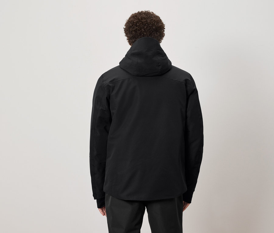 Mężczyzna o kręconych włosach stoi tyłem, ubrany w czarną kurtkę z kapturem.