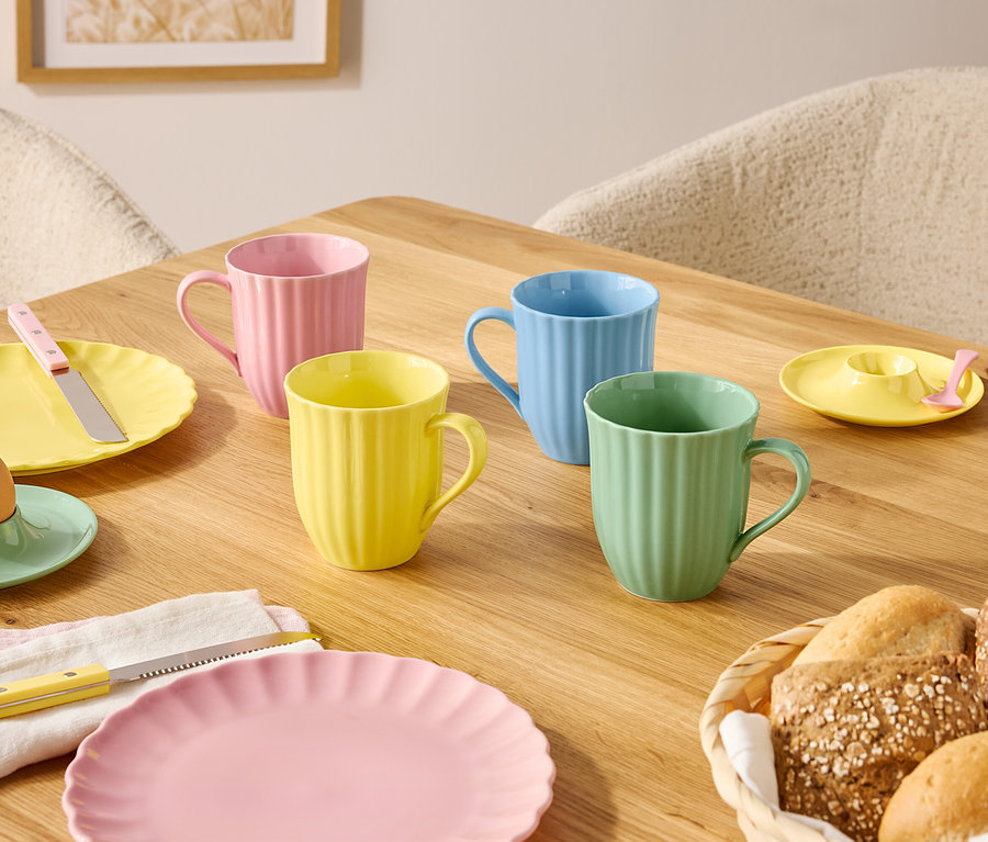 Na drewnianym stole stoją talerze i kubki w pastelowych kolorach oraz koszyk z pieczywem.