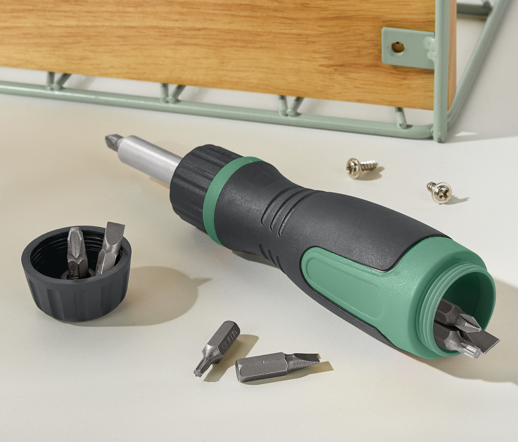 Czarno-zielony magnetyczny śrubokręt grzechotkowy leży obok pokrywki z bitami i dwóch śrub.