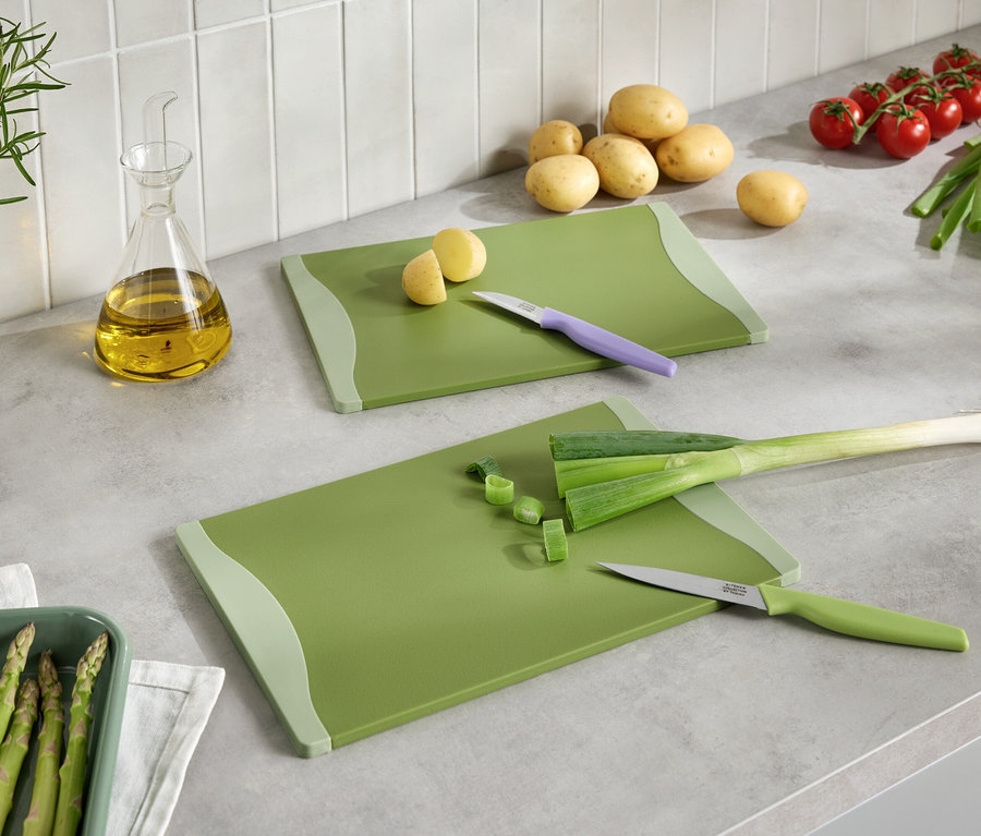 Dwie zielone deski do krojenia z pokrojonymi warzywami, noże, ziemniaki, pomidory, por, szparagi i olej na blacie kuchennym.