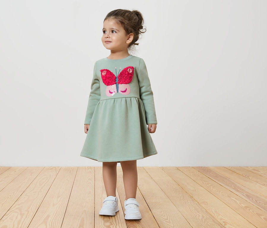 Dziewczynka w zielonej sukience z motylem stoi na drewnianej podłodze.