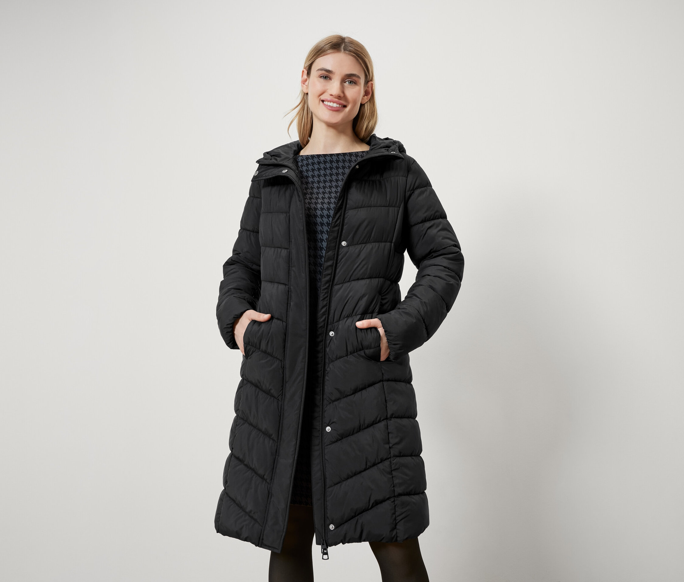 Kobieta o blond włosach stoi i ma na sobie długi, czarny płaszcz puchowy z kapturem.