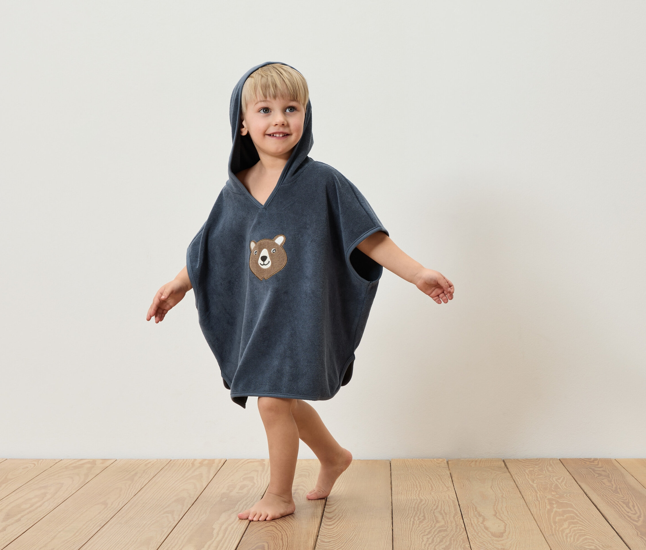 Chłopiec w niebieskim ponczo z kapturem i aplikacją misia stoi na drewnianej podłodze na tle białej ściany.