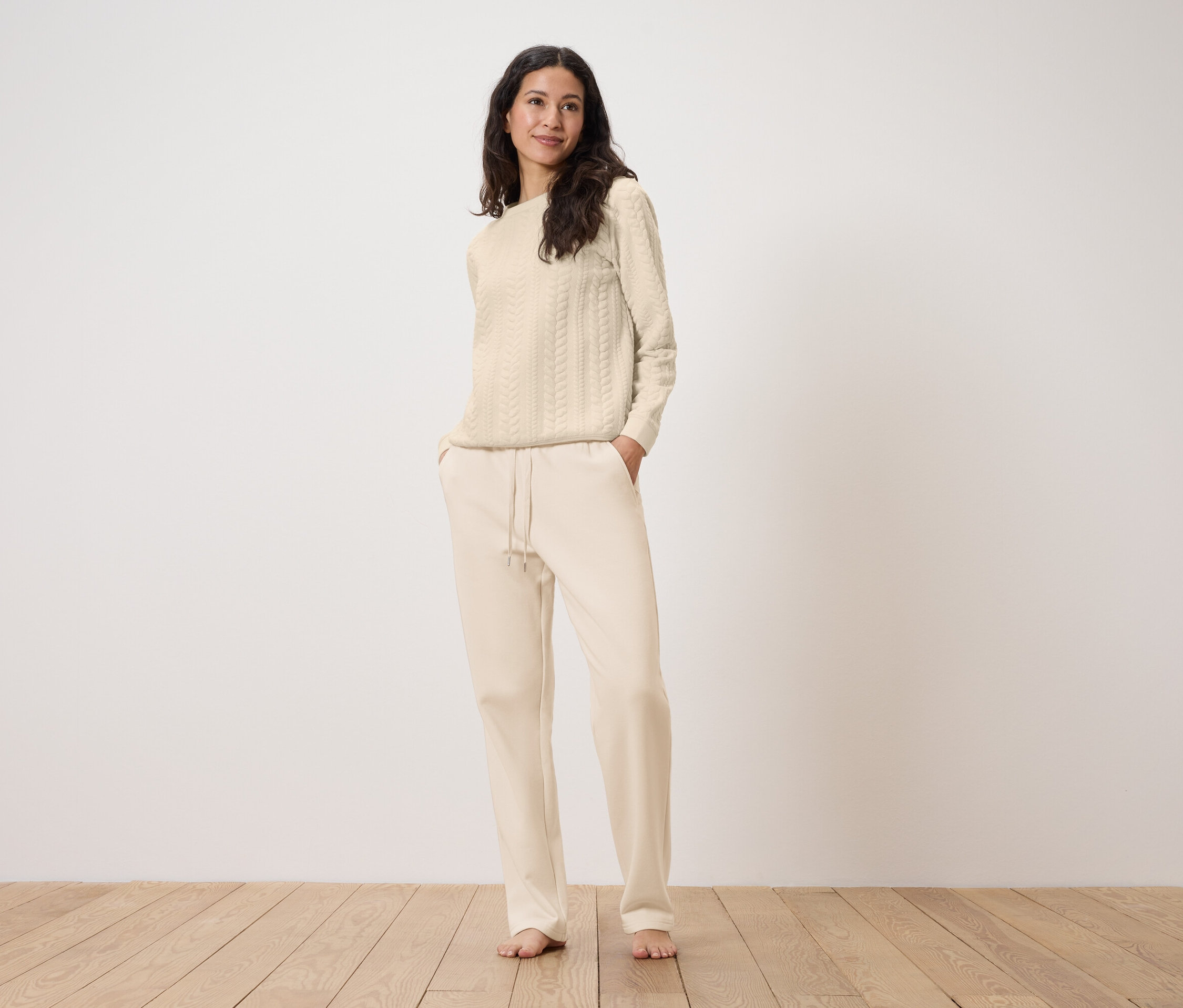 Kobieta stoi z rękami w kieszeniach kremowych spodni dresowych i ma na sobie kremowy sweter w warkocze.