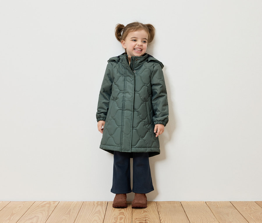 Uśmiechnięta dziewczynka z warkoczykami stoi na tle białej ściany, ubrana w zielony płaszcz, granatowe spodnie i brązowe buty.
