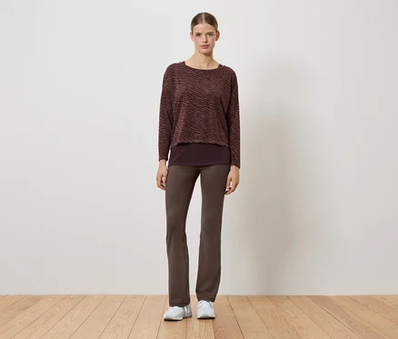 Kobieta w brązowym swetrze i spodniach stoi na drewnianej podłodze.