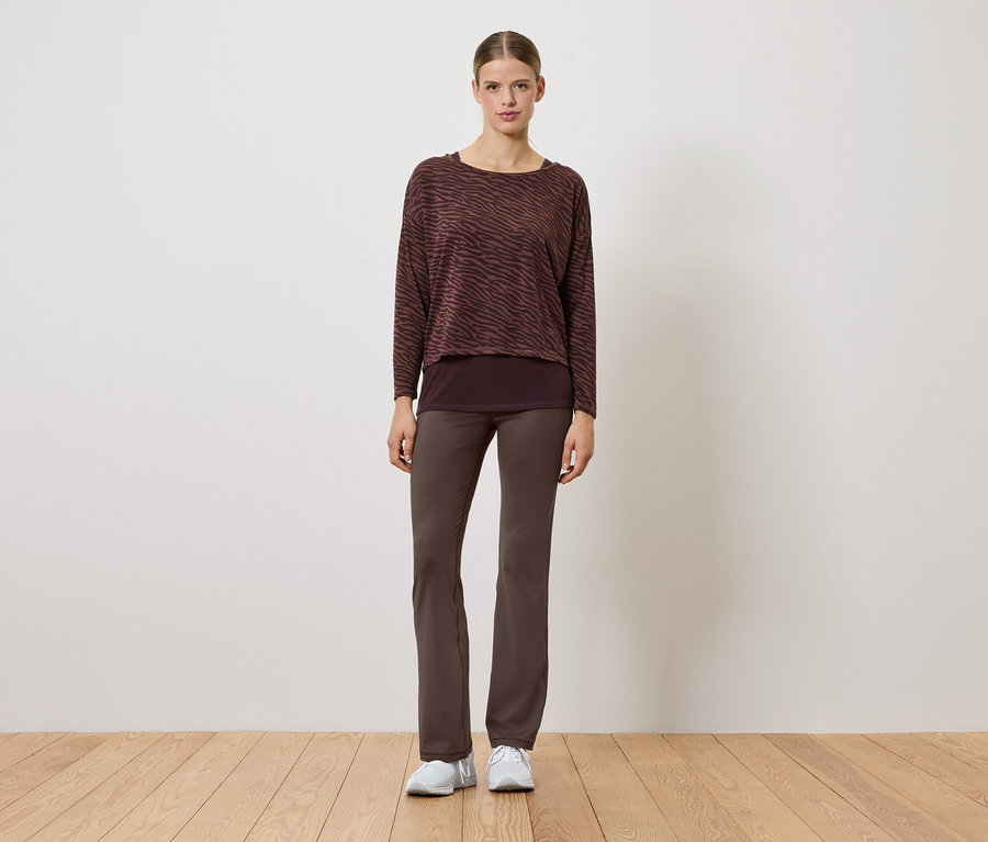 Kobieta w brązowym swetrze i spodniach stoi na drewnianej podłodze.