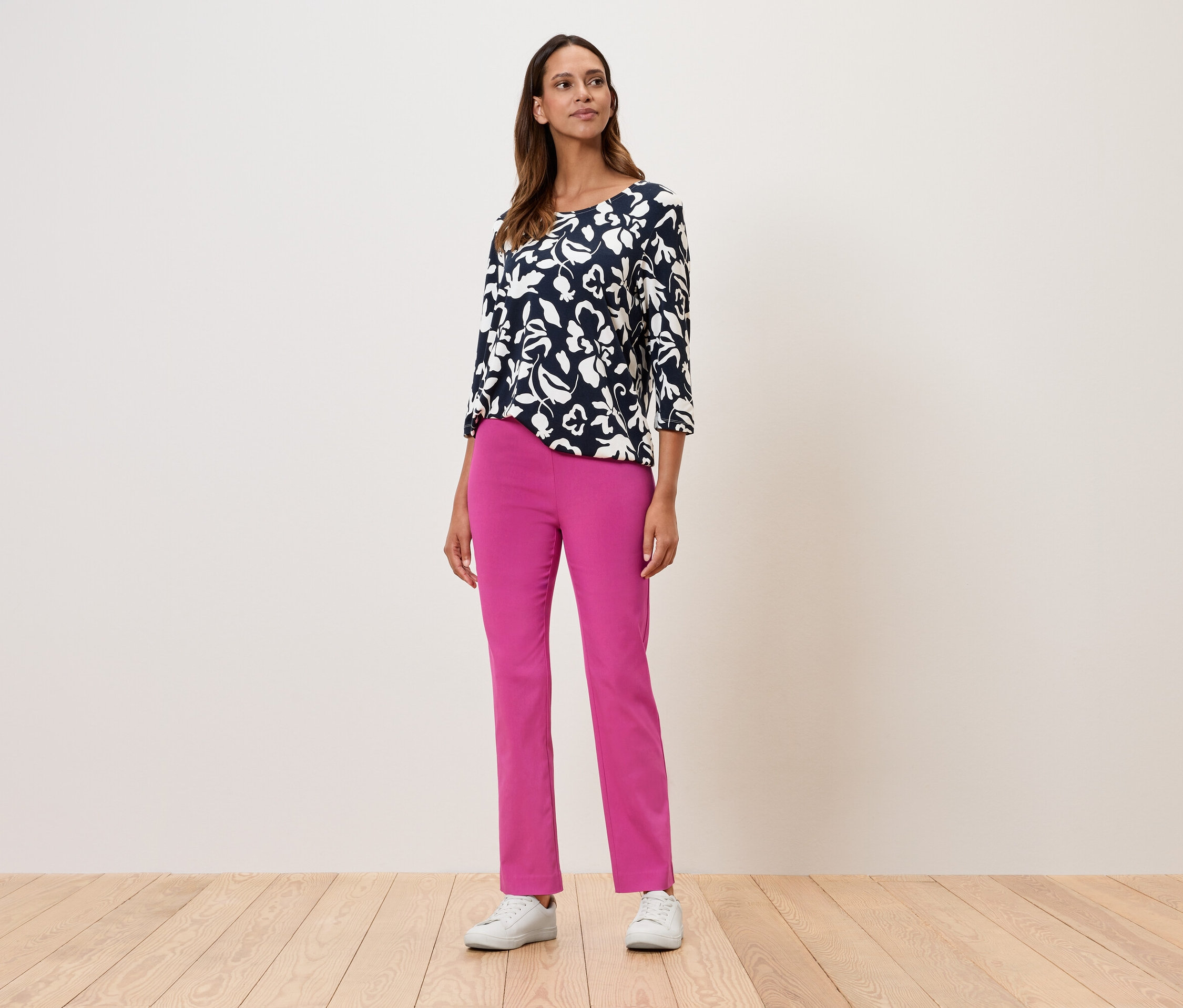Kobieta stoi i ma na sobie bluzkę w kwiaty z rękawami trzy czwarte, różowe spodnie i białe tenisówki.