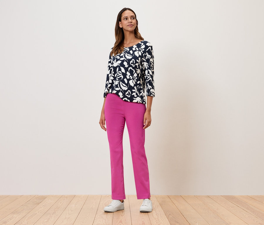 Kobieta stoi i ma na sobie bluzkę w kwiaty z rękawami trzy czwarte, różowe spodnie i białe tenisówki.