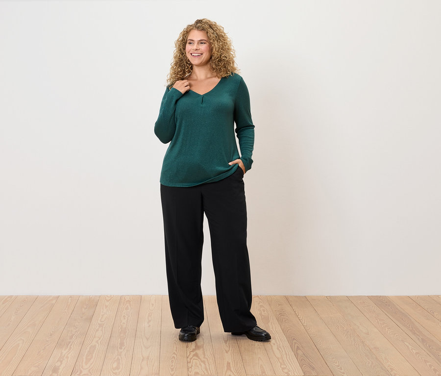Kobieta z kręconymi włosami pozuje w szmaragdowozielonym swetrze i czarnych spodniach.