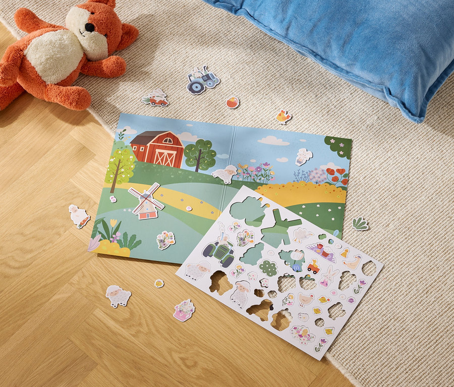 Pluszowy lis leży obok otwartej książki z ilustracją farmy i arkusza z naklejkami z farmą na dywanie.