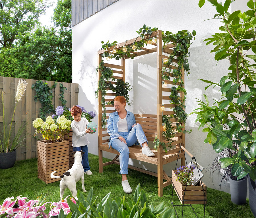 Kobieta siedzi na ławce ogrodowej z pergolą, podczas gdy dziecko bawi się piłką. Nad ławką wisi łańcuch świetlny LED „Żarówki”. W ogrodzie stoją 2 donice-kolumny na rośliny »Tinus« i skrzynka na zioła z ramką do przenoszenia „Elin”.