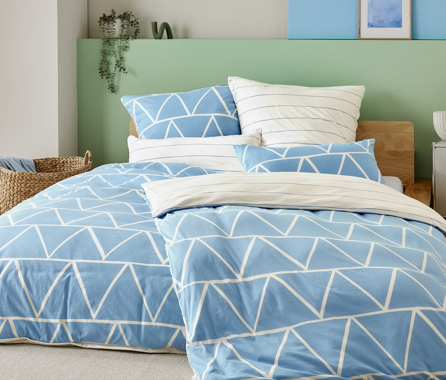 Łóżko z jasno niebieskim pokrowcem w białe trójkąty i poszewkami w paski, obok kosza wiklinowego i szafki nocnej z lampką.