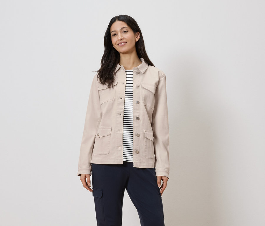 Kobieta ma na sobie beżową kurtkę nałożoną na koszulę w paski i granatowe spodnie.