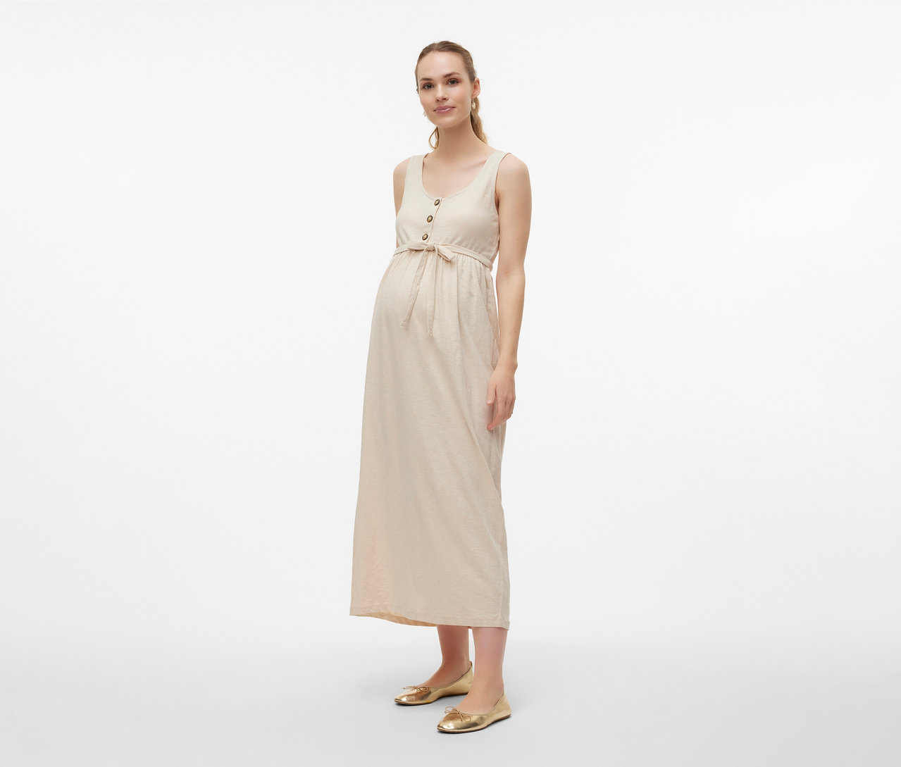 Kobieta w sukience ciążowej MAMALICIOUS „MLEVI LIA” z paskiem i złotych balerinach na jasnym tle.