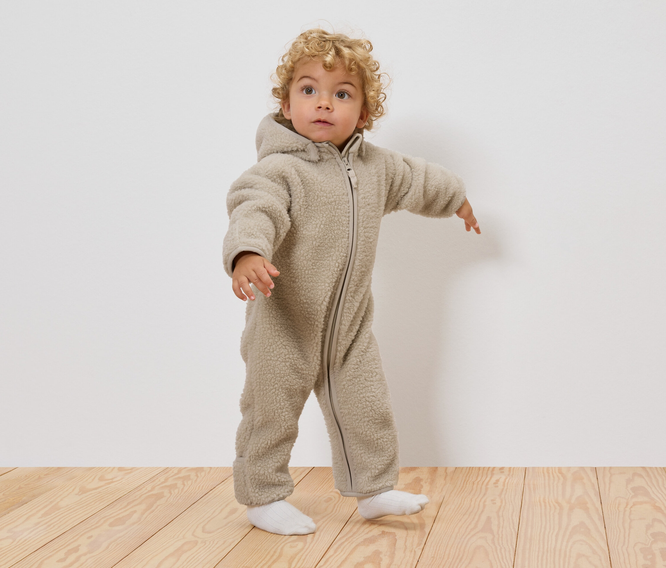 Małe dziecko z kręconymi blond włosami ma na sobie beżowy kombinezon typu teddy z kapturem i białe skarpetki, stojąc na drewnianej podłodze.