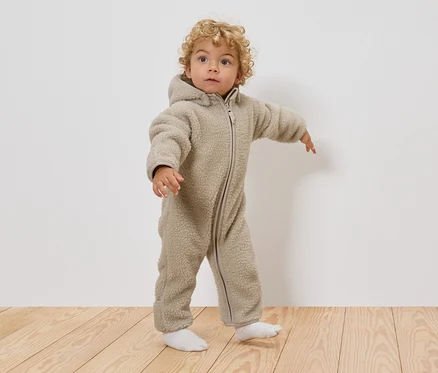 Małe dziecko z kręconymi blond włosami ma na sobie beżowy kombinezon typu teddy z kapturem i białe skarpetki, stojąc na drewnianej podłodze.