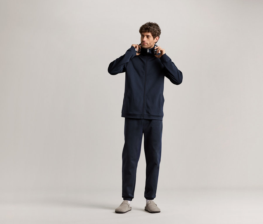 Mężczyzna stoi i trzyma słuchawki w ręku, ubrany w granatowy dres domowy.