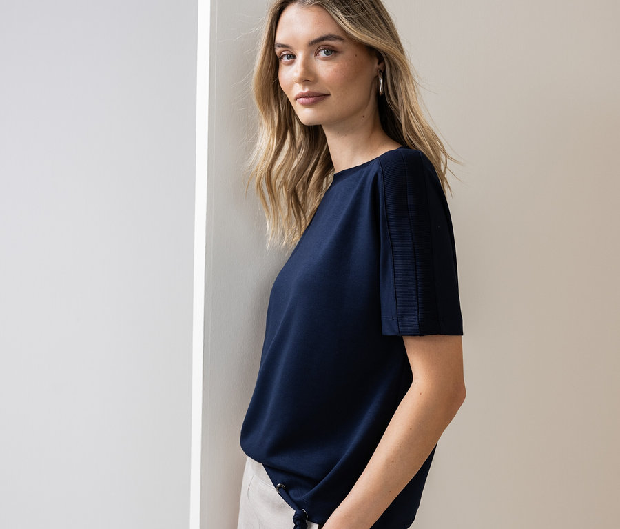 Kobieta opiera się o ścianę, ubrana w granatową koszulkę i jasne spodnie.