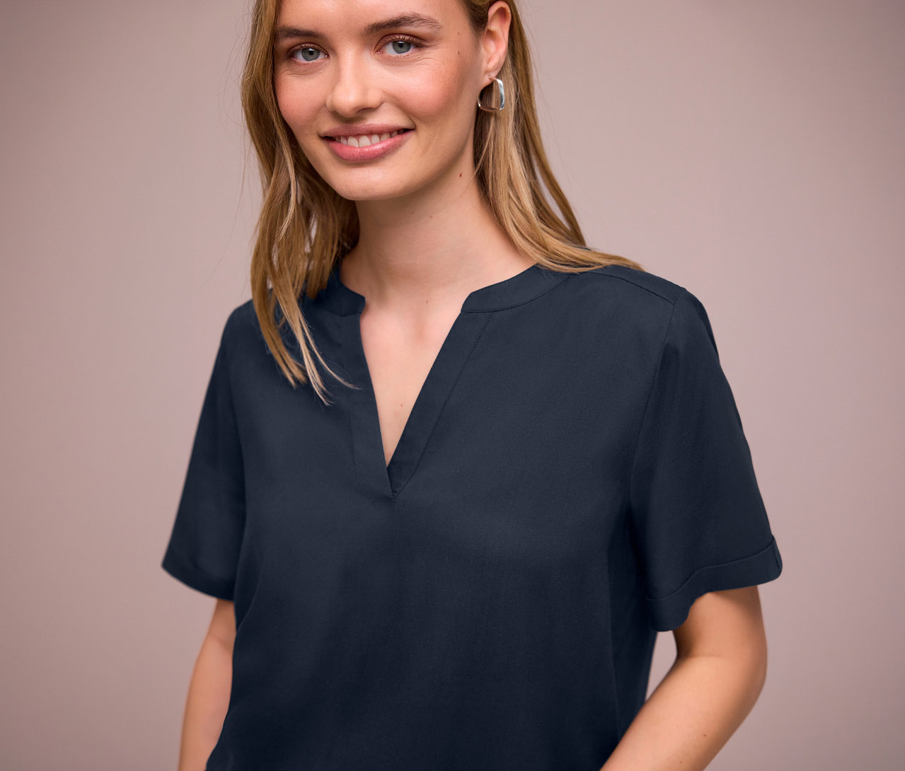 Kobieta ma na sobie granatową bluzkę z tkaniny Street One z dekoltem w szpic i srebrne kolczyki.